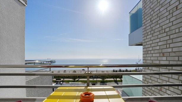 Uma varanda com uma mesa amarela, um objeto vermelho e vista para o mar e um cais.