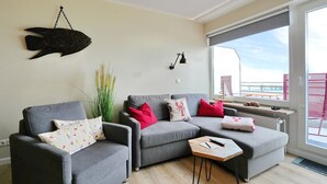 Uma sala de estar moderna com um sofá cinzento, uma pequena mesa de centro de madeira e uma grande janela com vista para o exterior.