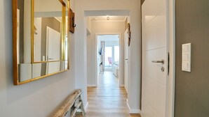 Um corredor com um banco de madeira, um espelho e uma porta que dá para um quarto com cama e janela.