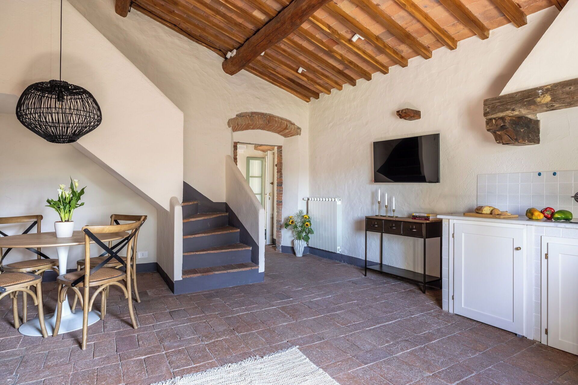 A rustic kitchen with a wooden staircase, a dining area with wicker chairs, and a TV mounted on the wall.