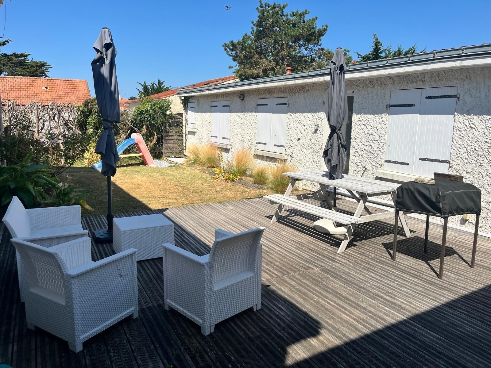 Een terras buiten met een houten dek, witte rieten meubels, een picknicktafel en parasols.