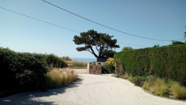 A gravel driveway leading to a gate, surrounded by greenery and a clear sky.