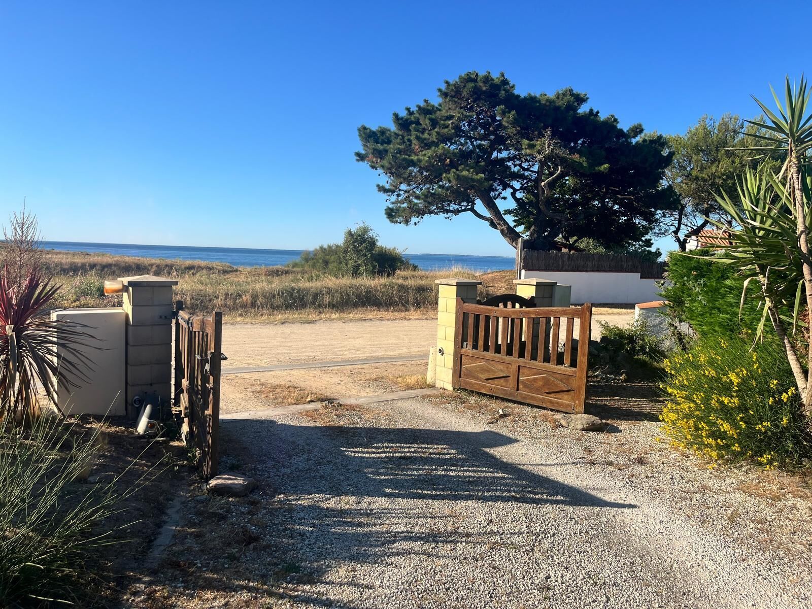 Une allée de gravier menant à une entrée fermée par un portail, d’où l’on a une vue sur la mer.