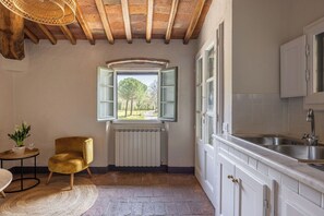 A kitchen with white cabinets, a stainless steel sink, a window with green shutters, a radiator, and a yellow chair. - Maison Badia a Passignano, Aleatico (Badia A Passignano)