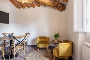 Ein Zimmer mit Holzboden, einem runden Tisch mit Stühlen, einem kleinen Tisch mit Blumenvase und Glas sowie einem Heizkörper.