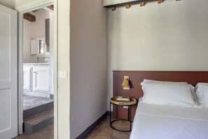 A bedroom with a bed, a bedside table with books, a wall-mounted lamp, and a door leading to a bathroom. - Maison Badia a Passignano, Aleatico (Badia A Passignano)