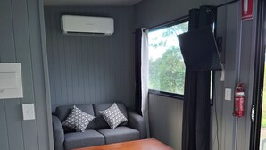 A room with a grey sofa, patterned cushions, a wooden coffee table, a window with curtains, an air conditioning unit, and a fire extinguisher. - Gunabul Homestead & Golf Course (Gympie)