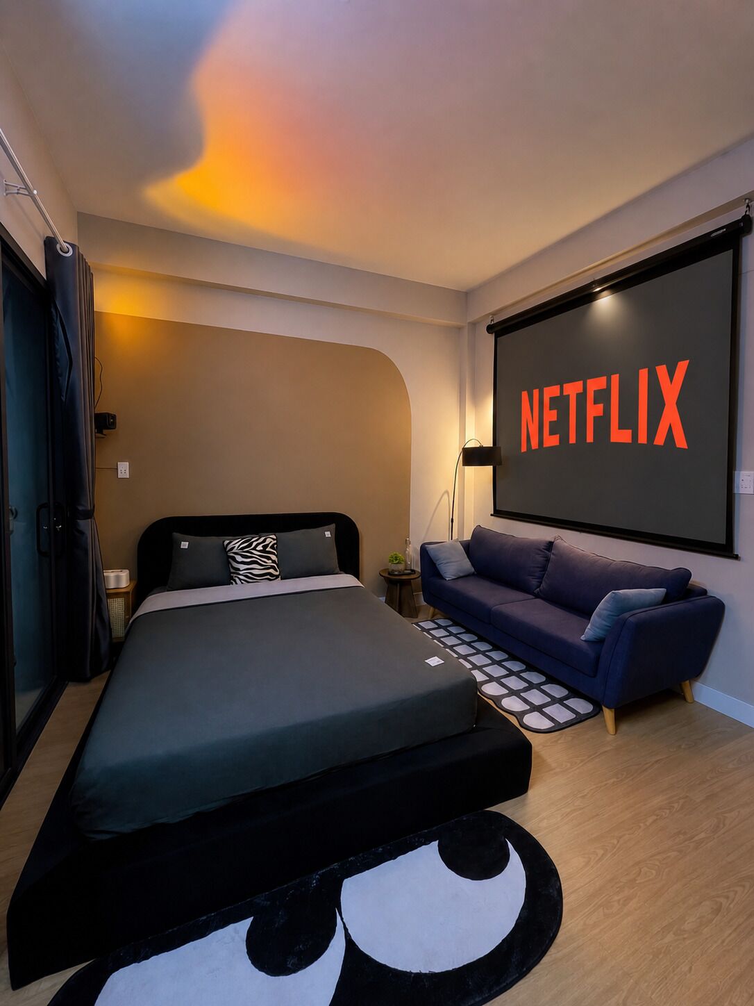 Une chambre moderne avec un lit, un canapé, un écran fixé au mur affichant le logo « NETFLIX », et une petite table d’appoint.