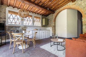 A rustic interior with a stone wall, wooden ceiling, a dining area with wicker chairs, a kitchen with white cabinets, and a cozy seating area with a sofa. - Maison Badia a Passignano, Canina Nera (Badia A Passignano)