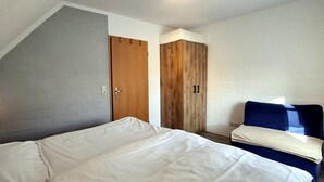 A bedroom with a sloped ceiling, a bed with white bedding, a wooden wardrobe, and a blue chair. - Appartmenthaus Nolting Apartment 3 Nolting: 40 m² with terrace and WLAN - (Werdum)