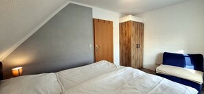 Ein Schlafzimmer mit schrägverdeckter Decke, einem Bett mit weißer Bettwäsche, einem Holzschrank und einem blauen Sessel.