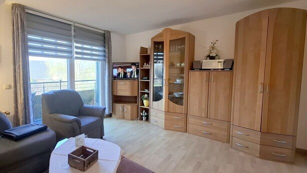 A living room with a sofa, a chair, a table with a box, a cabinet with glass doors, and a window with blinds. - Appartmenthaus Nolting Apartment 3 Nolting: 40 m² with terrace and WLAN - (Werdum)