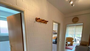 En hall med en vit tegelvägg, en trädörr, en liten trähylla, en klocka och ett fönster med persienner.