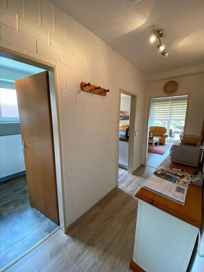 Un pasillo con pared de ladrillo blanco, una puerta de madera, un pequeño estante de madera, un reloj y una ventana con persianas.