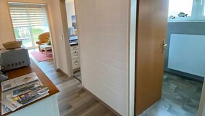 A hallway with a wooden floor, a white wall with a brick pattern, a door with a round handle, and a ceiling light fixture. - Appartmenthaus Nolting Apartment 2 Nolting with terrace & WLAN in Werdum - (Werdum)