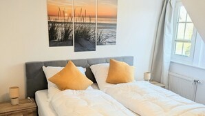 A double bed with white bedding and two pillows, a bedside table with a lamp, and a window with curtains. - Vacation home Am Kurzentrum 26 No. 3 (Wittmund)