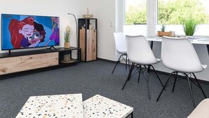 A modern room with a TV, a dining area with chairs, a window with a view, and a small plant. - Deich in Sicht - Holiday apartment "Deich in Sicht" (Neuharlingersiel)