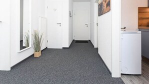 A hallway with white walls, a grey carpet, a potted plant, a door, and a small white appliance. - Deich in Sicht - Holiday apartment "Deich in Sicht" (Neuharlingersiel)