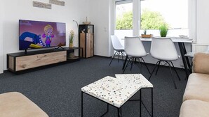 A modern living room with a TV, a dining area with chairs, and a window with a view. - Deich in Sicht - Holiday apartment "Deich in Sicht" (Neuharlingersiel)