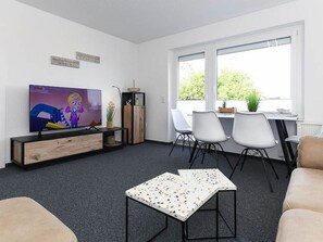 A modern living room with a TV, a dining area with chairs, and a window with a view. - Deich in Sicht - Holiday apartment "Deich in Sicht" (Neuharlingersiel)