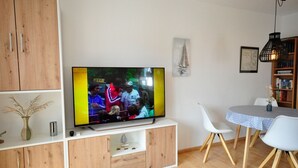 A living room with a flat-screen TV, wooden cabinets, a dining table with chairs, and a bookshelf. - Wohnpark Legewarfen 2/3 - Holiday apartment Wohnpark Legewarfen 2/2 (Werdum)