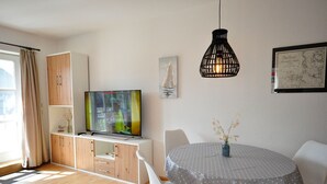 A modern living room with a TV, wooden cabinets, a dining table with chairs, a hanging light fixture, and a map on the wall. - Wohnpark Legewarfen 2/3 - Holiday apartment Wohnpark Legewarfen 2/2 (Werdum)