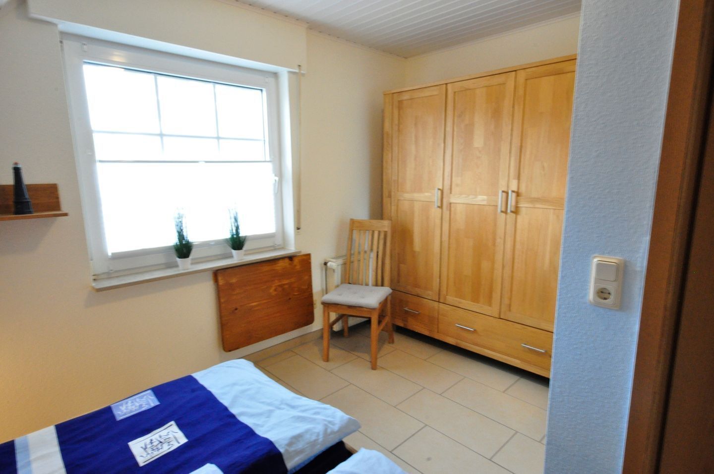 Un dormitorio con cama, armario de madera, silla, ventana con plantas y radiador.