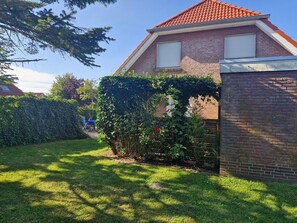 A residential building with a brick facade, a red-tiled roof, and a garden area with a hedge and a small tree. - Vacation home Uferstraße 2 F - Holiday home Uferglück Uferstraße 2 F (Wittmund)