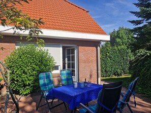 A patio with a table and chairs, a brick building with a red roof, and green foliage. - Vacation home Uferstraße 2 F - Holiday home Uferglück Uferstraße 2 F (Wittmund)