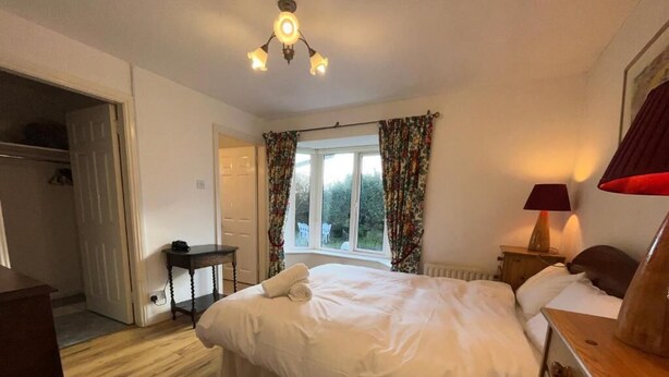 Een slaapkamer met een bed, nachtkastje, lamp, raam met gordijnen en een deur naar een andere kamer.