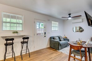Ein Zimmer mit einer Tür, zwei Barhockern, einer Couch, einem Esstisch mit Stühlen, einem Deckenventilator und einer wandmontierten Klimaanlage.