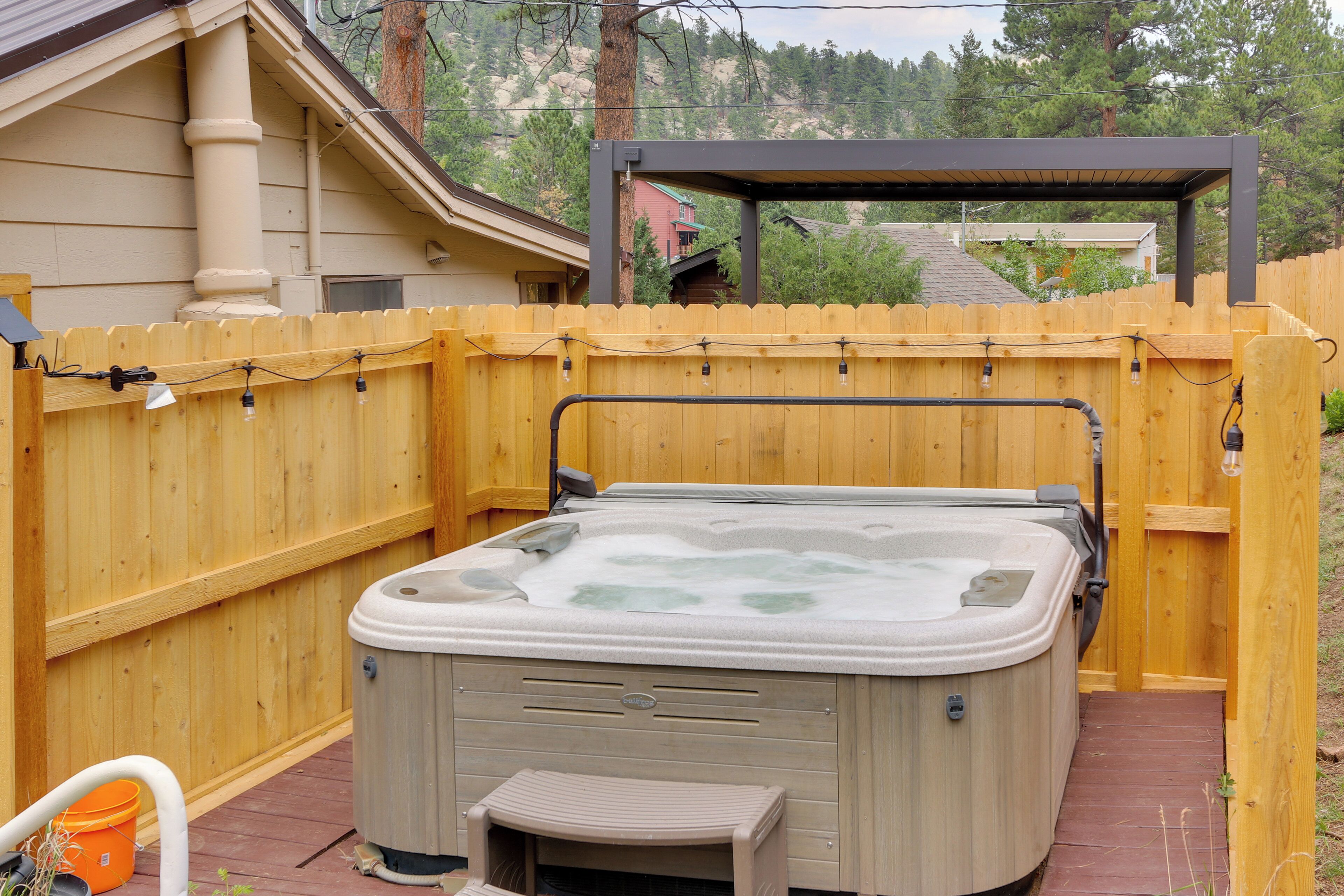 A hot tub in a backyard with a wooden fence, a pergola, and a view of a neighboring house.
