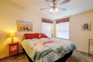 Un dormitorio con cama, dos ventanas con cortinas, ventilador de techo y decoración en la pared.