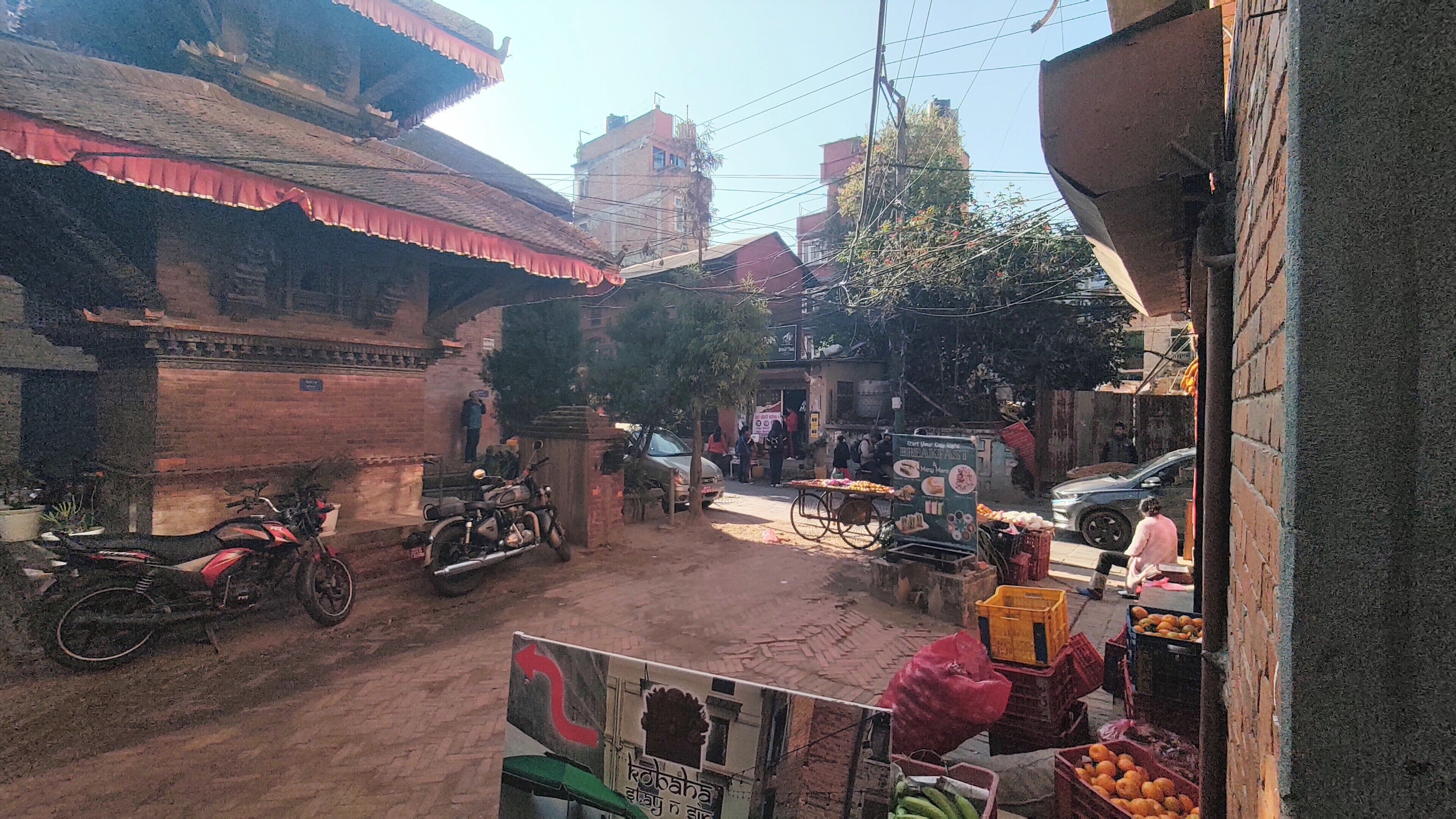 Una escena callejera con edificios, una motocicleta, una bicicleta y un carrito de vendedor con frutas.