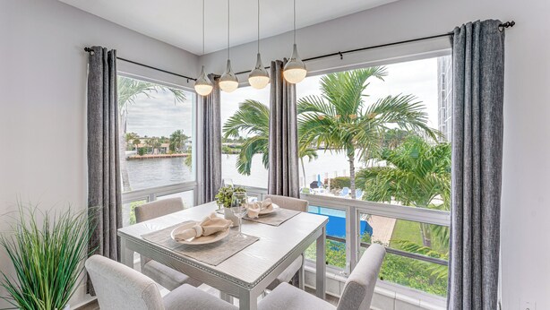 Un comedor con una mesa dispuesta para cuatro personas, ventanas amplias con cortinas y vistas a palmeras y una masa de agua.