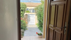 An open door leading to a tiled patio with a red mat, a wooden door with glass panels, and a view of a house with a tiled roof and greenery.