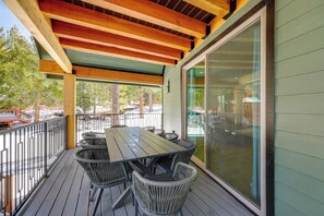 A covered outdoor patio with a wooden table and wicker chairs, a glass door, and a view of a snowy landscape. - Waterfront Luxury! Brand-New Big Bear Lake House (Big Bear Lake)