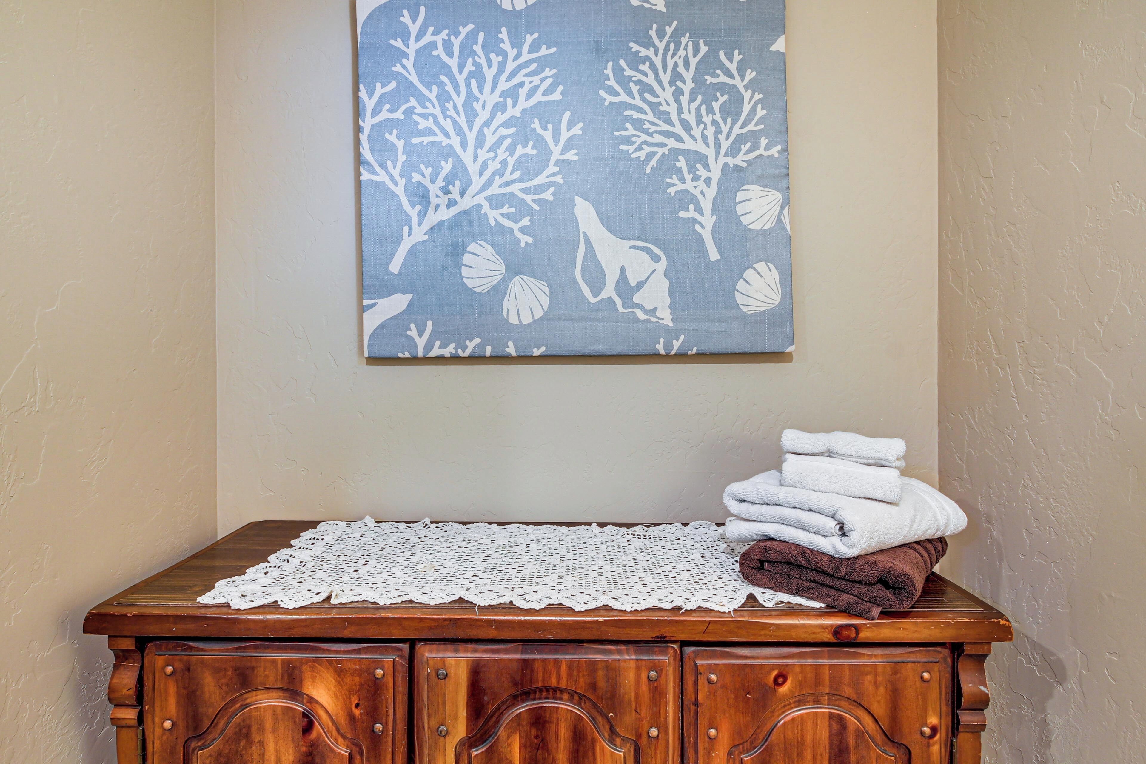 A wooden dresser with a white lace doily, a blue painting with white tree branches and leaves, and a stack of towels.