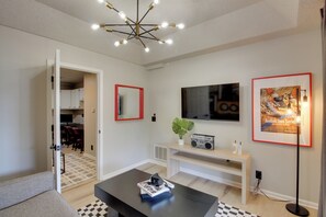 Un salon moderne avec une table basse noire, un téléviseur fixé au mur, un tableau encadré et une plante posée sur une console blanche.