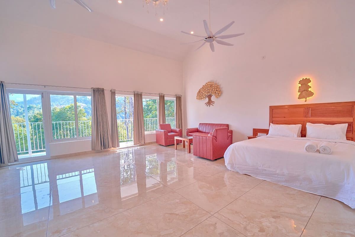 A spacious bedroom with a large bed, a ceiling fan, a nightstand, and a view of the outdoors through large windows.