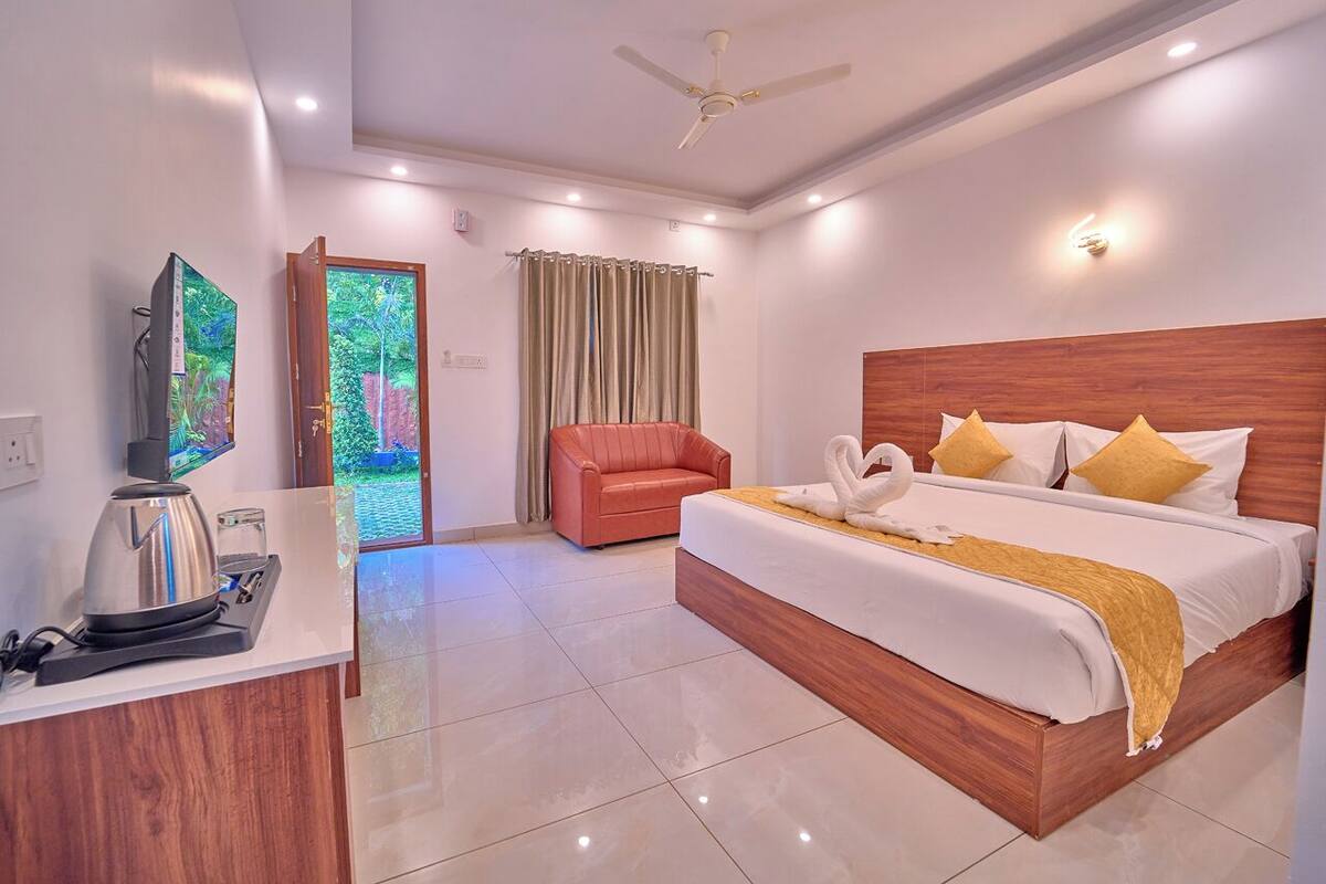 A hotel room with a bed, a desk with a kettle, a chair, and a view of greenery outside.