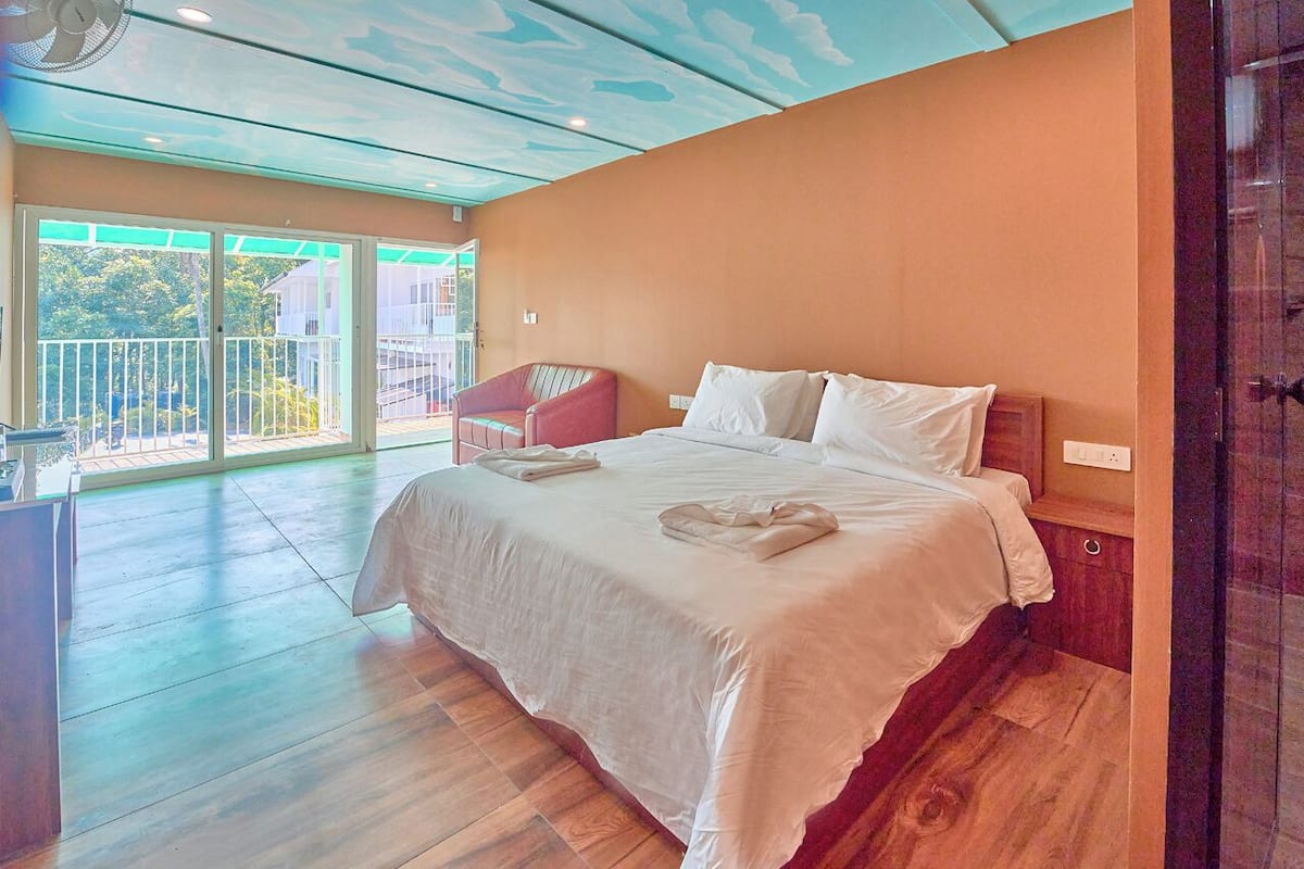 A bedroom with a large bed, a chair, a balcony with a railing, and a ceiling with a cloud design.
