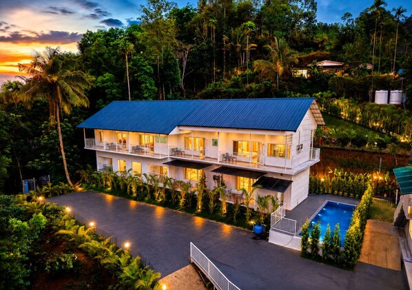 A two-story building with a flat roof, surrounded by lush greenery and a swimming pool. - Ice Cliff Munnar by VOYE HOMES (Udumbanchola)