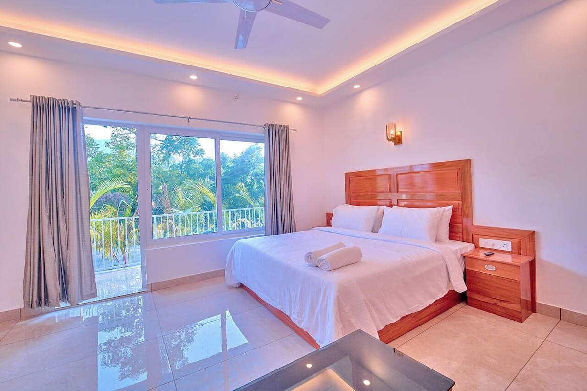 A modern hotel room with a large bed, a ceiling fan, a nightstand, and a balcony with a view of greenery.