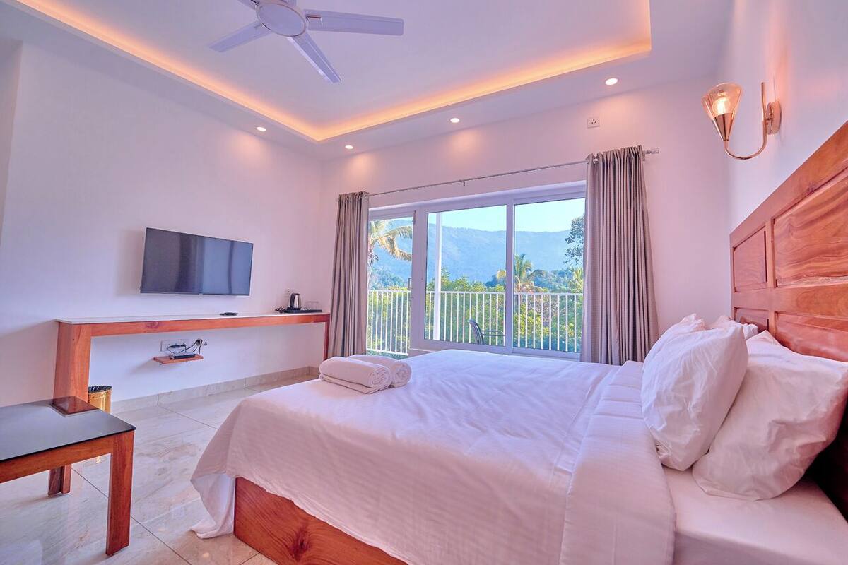 A hotel room with a bed, a TV, a desk, a ceiling fan, and a balcony with a view.