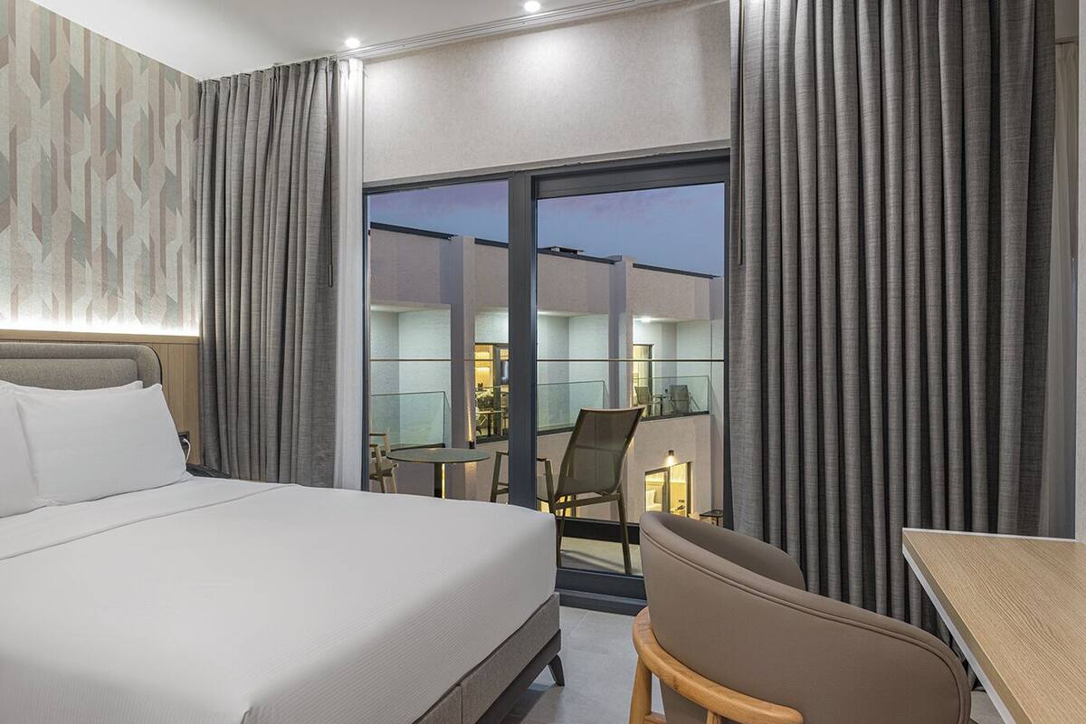 A hotel room with a bed, a desk, a chair, and a balcony with a glass door.