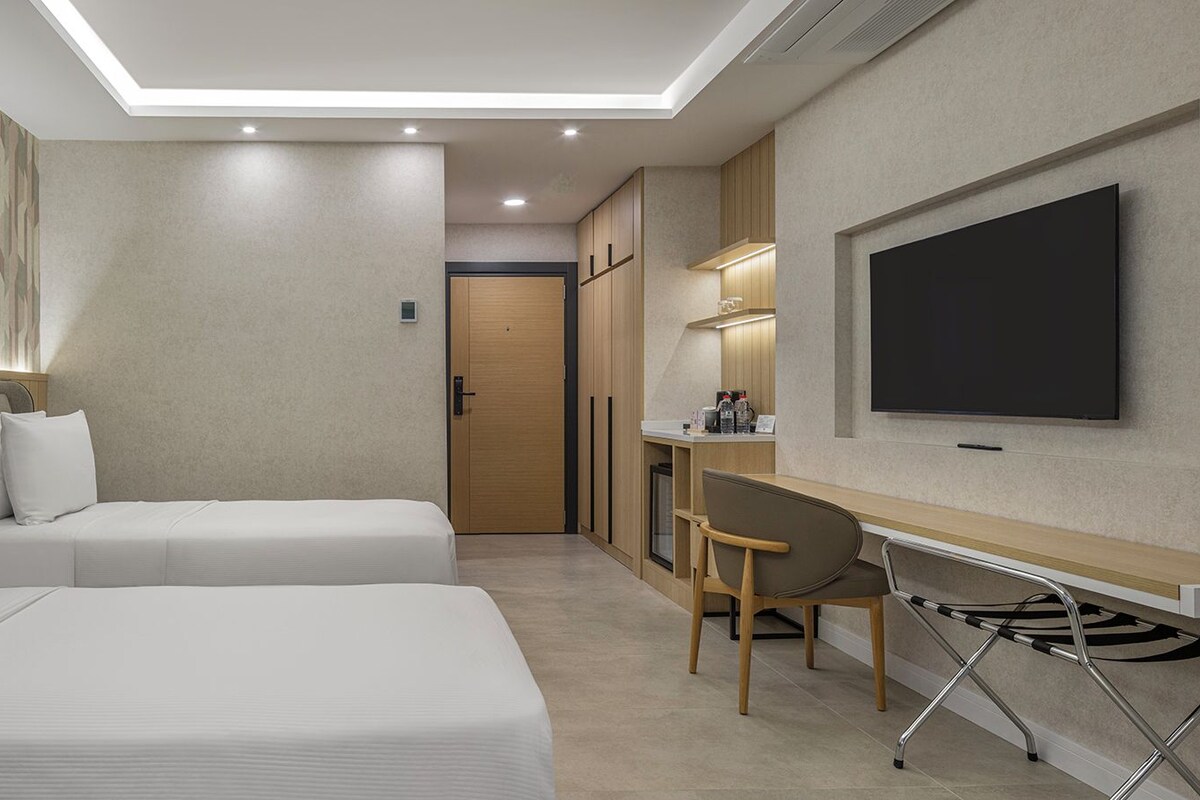 A hotel room with two beds, a desk, a chair, a TV, and a door.