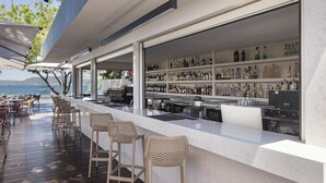 Un área de bar al aire libre con vista al mar, taburetes de mimbre y una variedad de botellas en estantes.