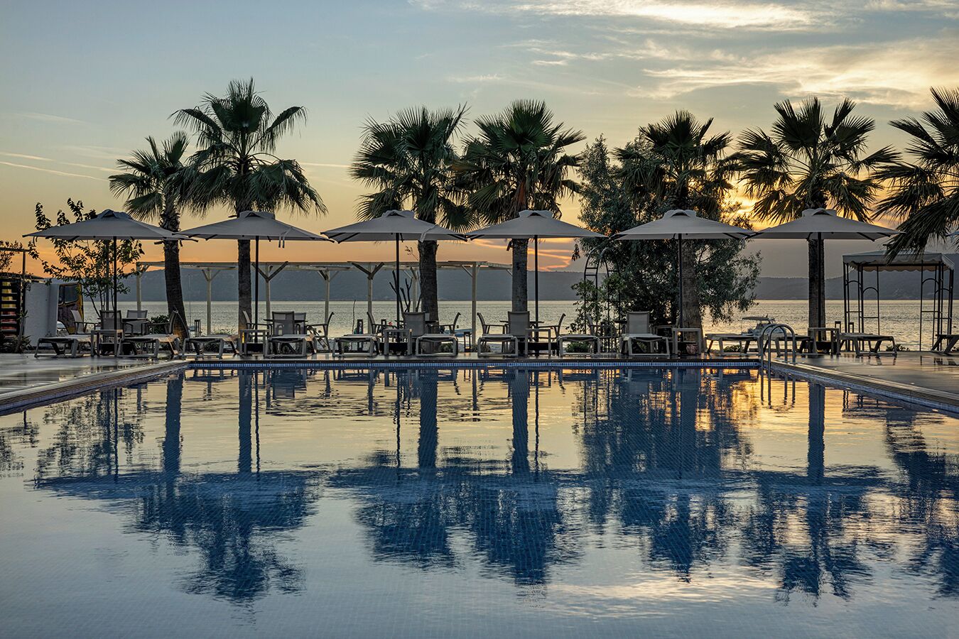 A pool with lounge chairs and umbrellas, palm trees, and a view of the sea at sunset.