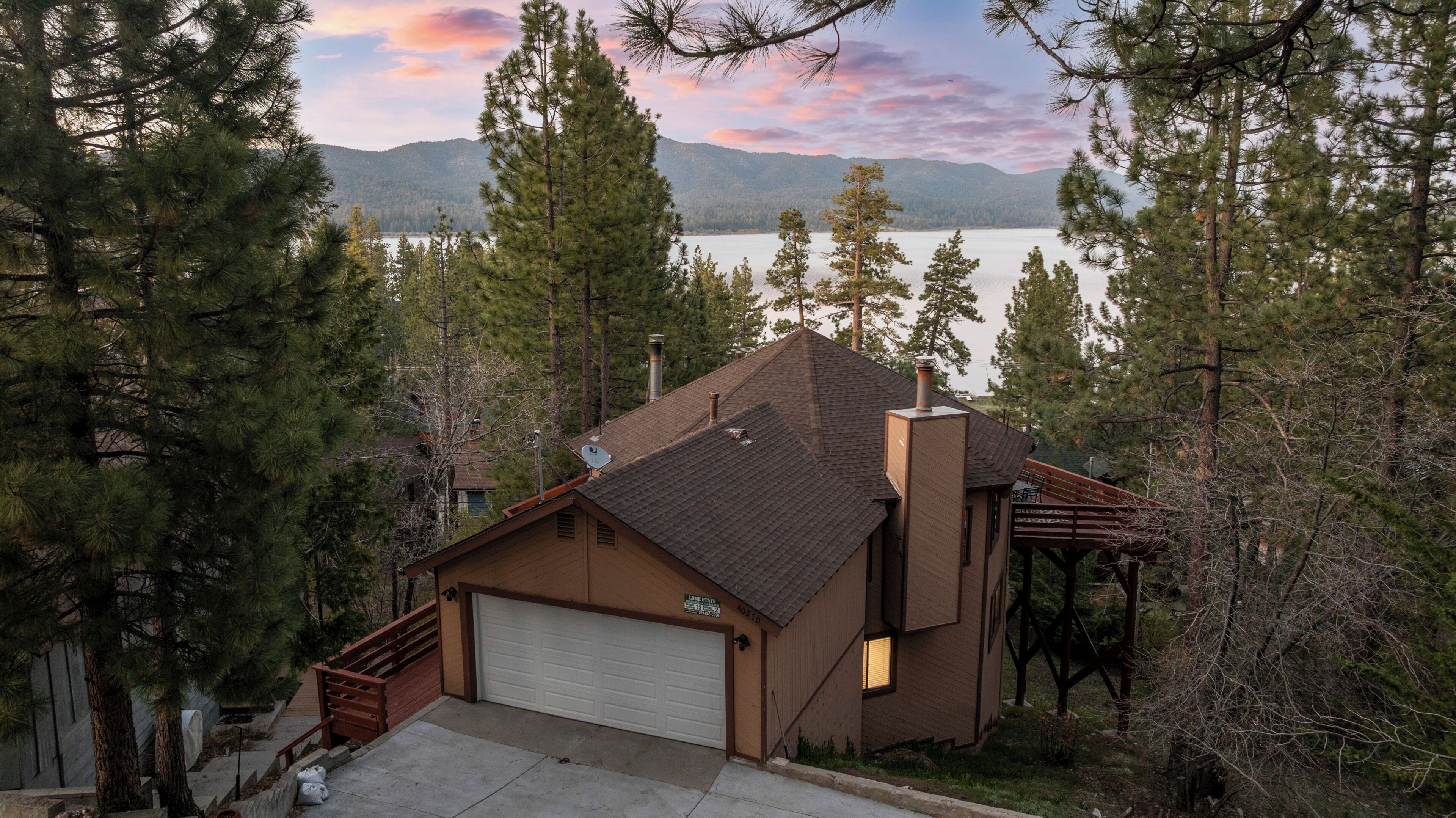 Une maison à deux étages avec un garage, entourée de grands pins et offrant une vue sur un lac.